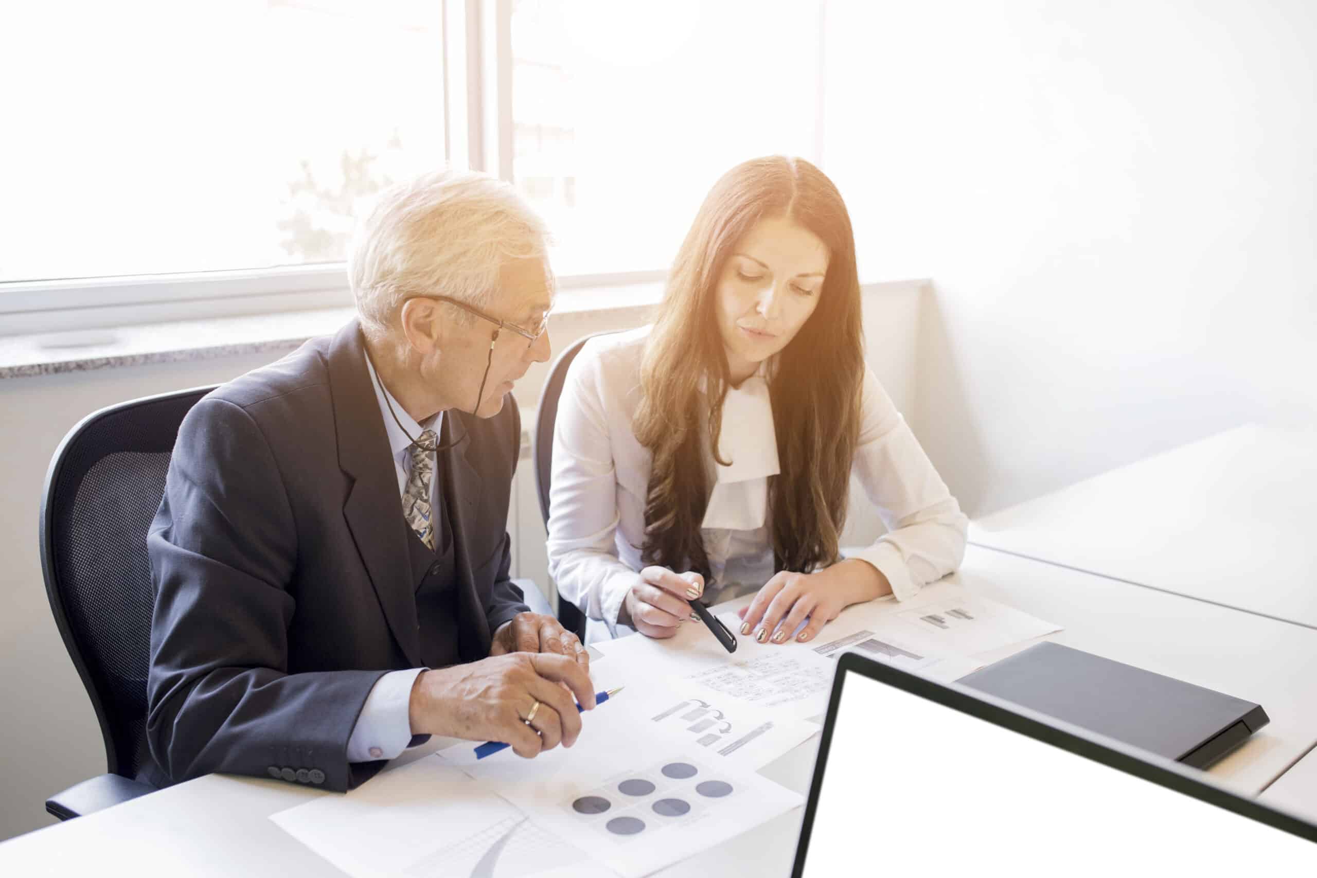 Imagem ilustrativa para as Big Four de Auditoria - Homem branco de cabelos brancos, óculos de grau e terno conversa com mulher branca de cabelos castanhos e camisa branca. Ambos estão sentados à mesa, onde há papéis com gráficos e um computador.